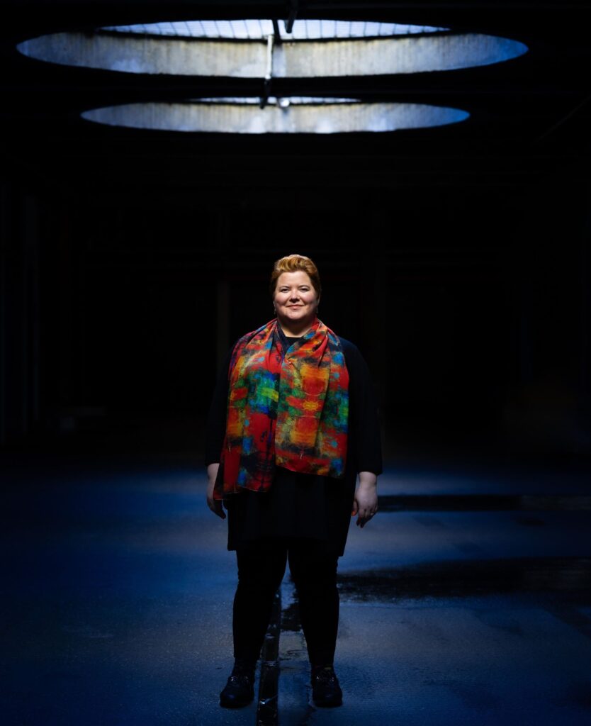 Woman standing, lit from above, in a dark space that looks a bit like a stage