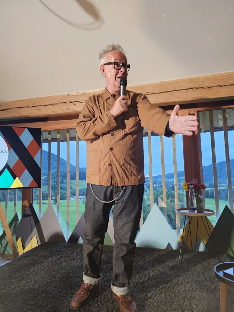 Mark, a white man wearing a brown utility jacket and jeans with a chain on them, holding a mic and gesturing at the crowd