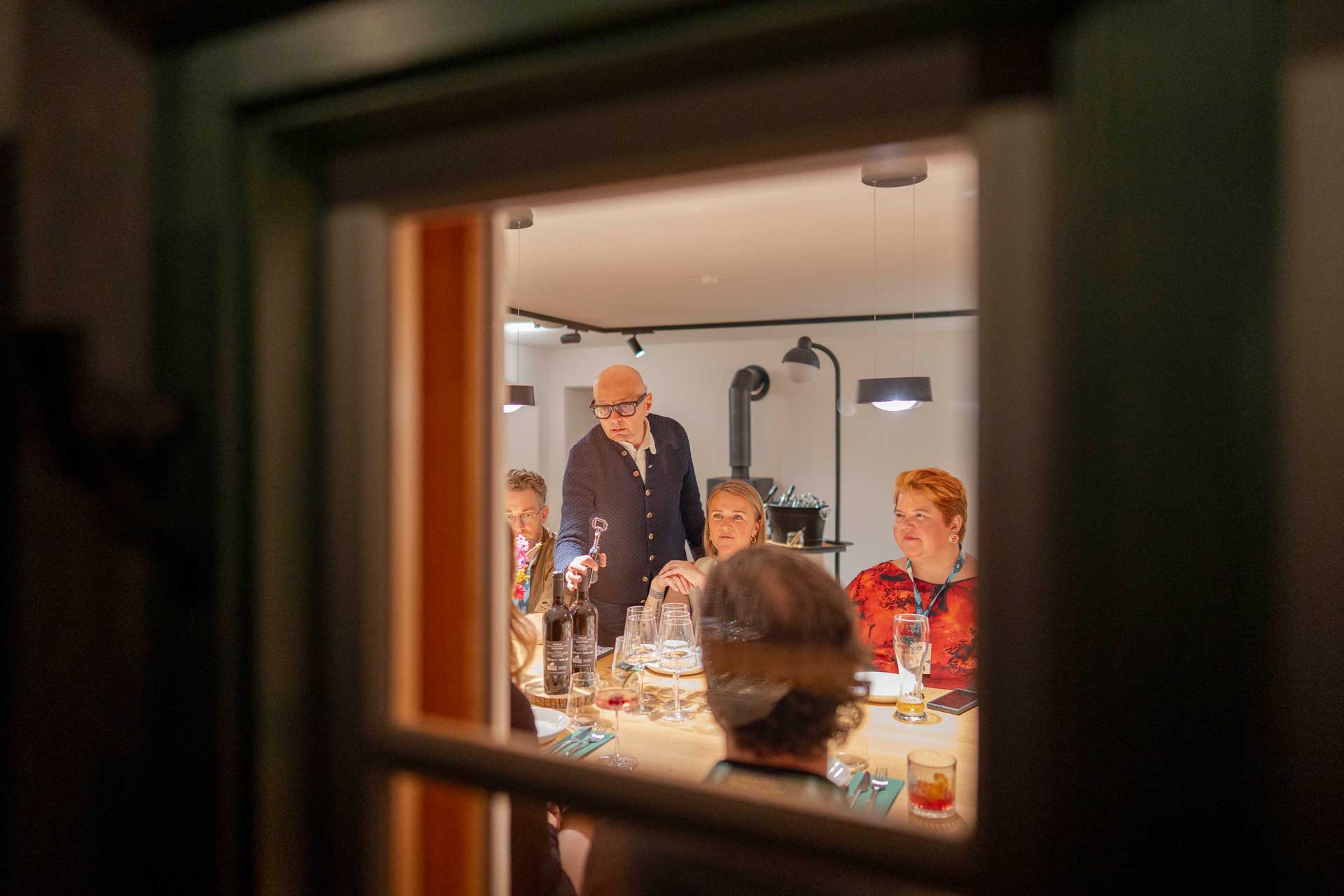 Looking into a warmly lit room through a window, where people are sat around a table having fun
