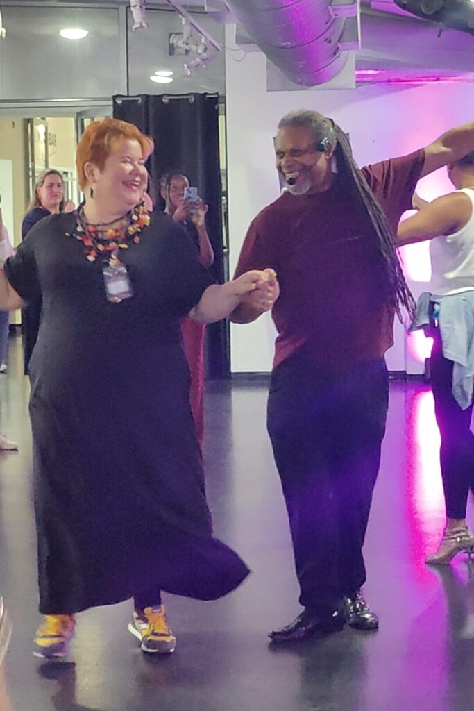 A woman in a black dress dances with a man with long dreadlocks and they're both smiling