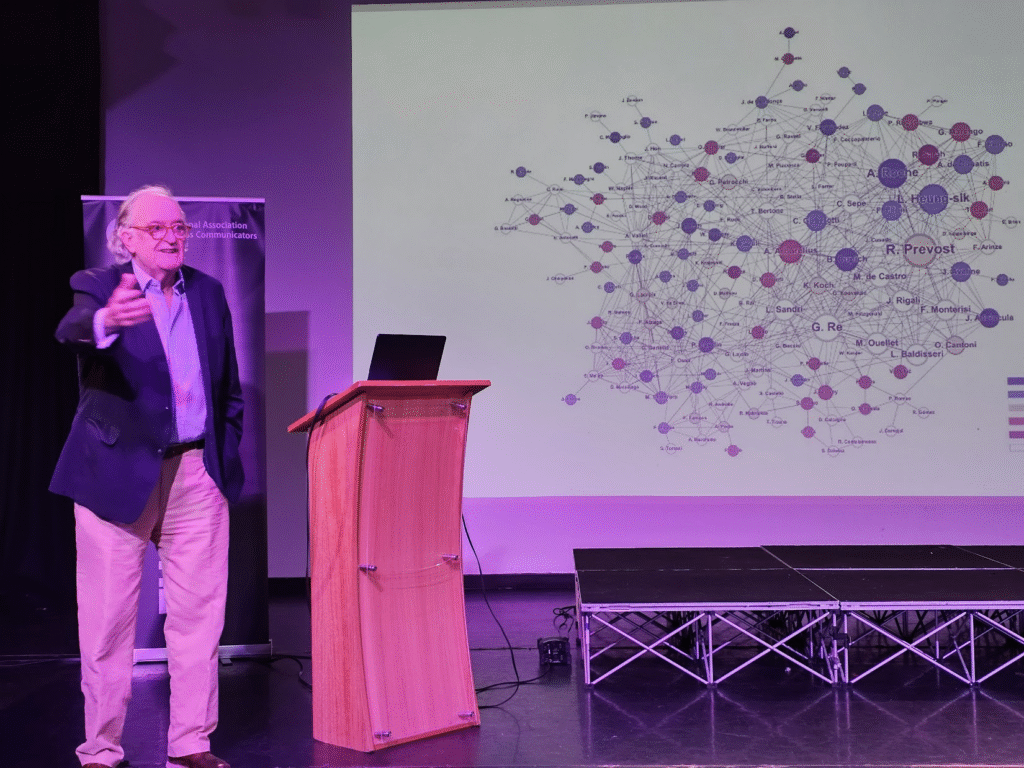 A man stands on stage showing a slide that has lots of connections and a large circle saying R Provost