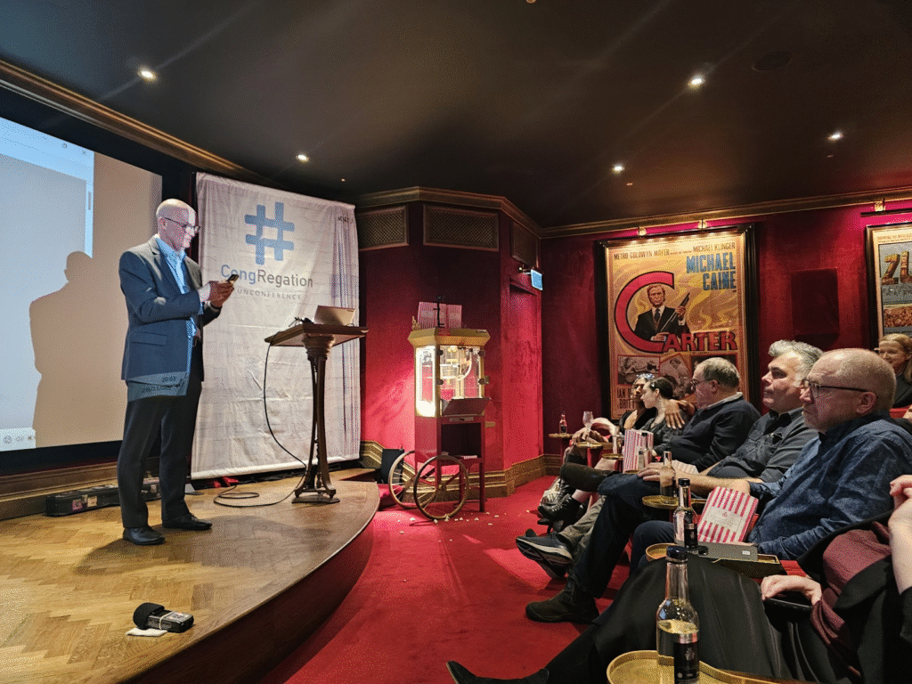 Man standing on a stage in a vintage cinema setting. There's a popcorn machine at the back and people sitting in the audience