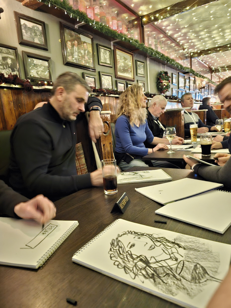 A group of people sitting around a table sketching. There's a drawing on the table of a woman with curly hair and a glass of wine