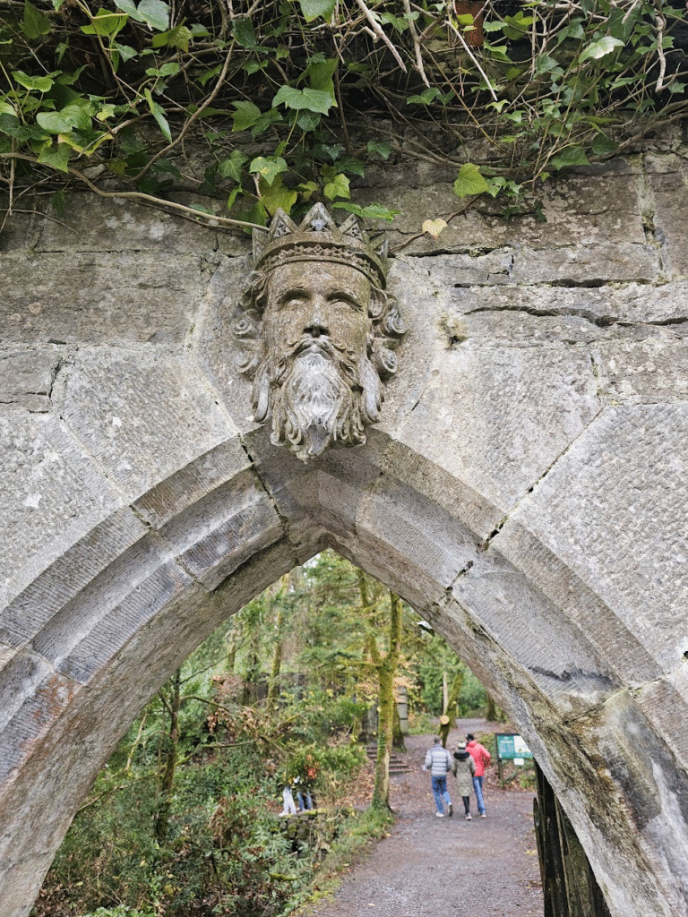 Stone arch with a carving of a bearded king, through the archway you can see three people walking in the distance