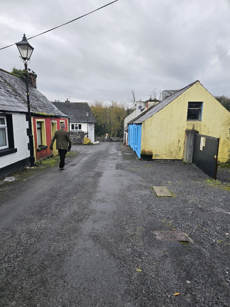 Down a back street in an Irish village, following a man wearing a tweed jacket. The houses look small and old. It's overcast.